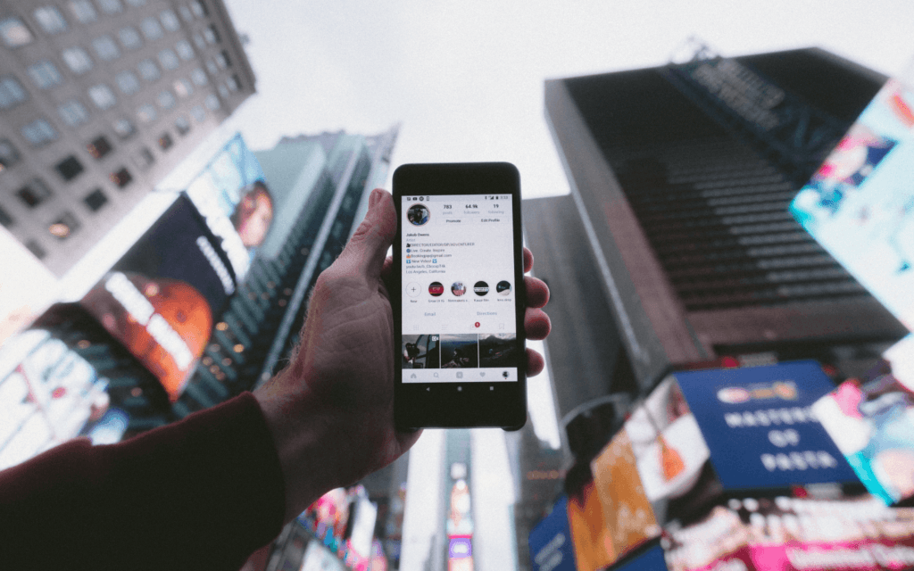 "A smartphone displaying an Instagram profile held against a blurred city backdrop with bright billboards, highlighting the reach of social media analyzed by an Instagram Insights Report."
