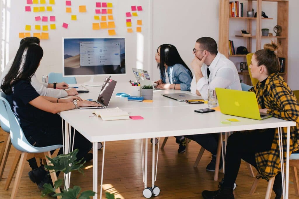 A team of professionals collaborating around a table with laptops, viewing a presentation on a screen, representing the use of AI tools for marketing to analyze data and optimize campaigns.