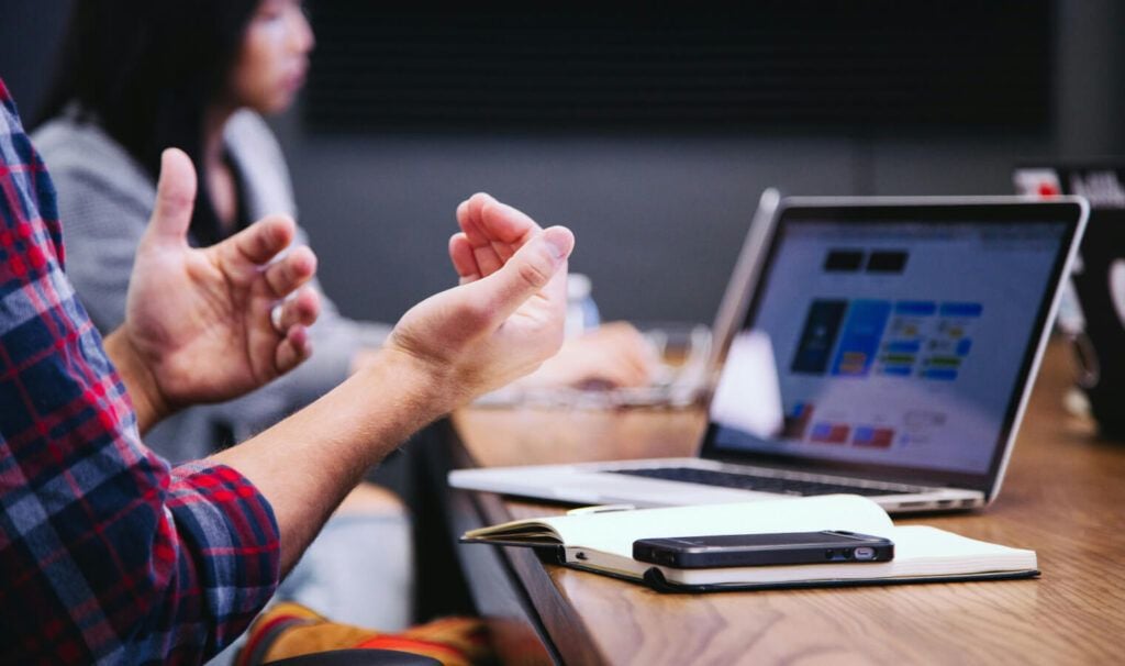 A person gesturing in a meeting with a blurred background, a laptop with an SEO dashboard on screen, and work essentials on a table, illustrating an in-depth SEO strategy session.