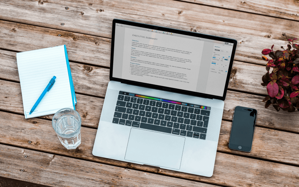 An overhead view of a work setup with a laptop displaying a document on custom reporting tools, accompanied by a notepad, pen, smartphone, and a glass of water on a wooden table.