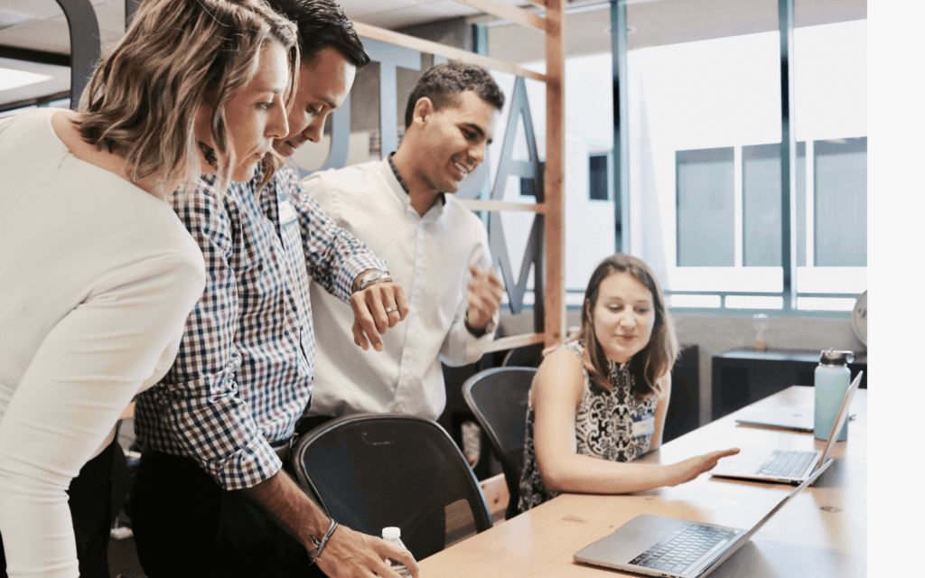 Four digital agency team members collaborating around a laptop, discussing strategies as part of their Digital Agency Workflow in a contemporary office environment.