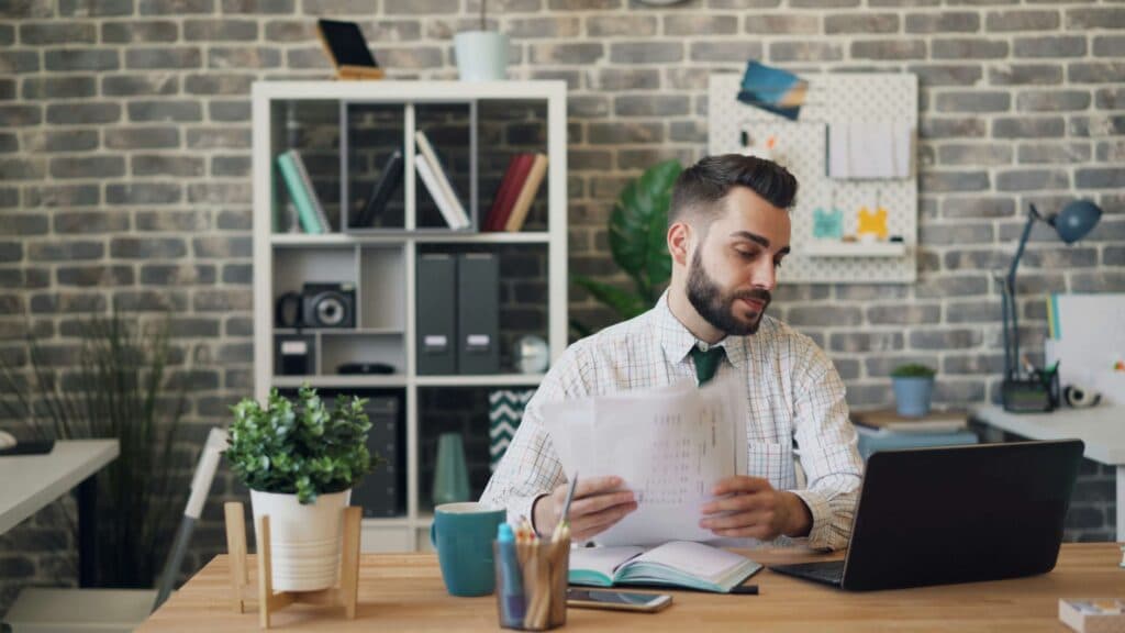 A man sitting at a desk with a laptop and documents, analyzing data and reports, representing the use of AI tools for marketing to generate insights and optimize strategies.