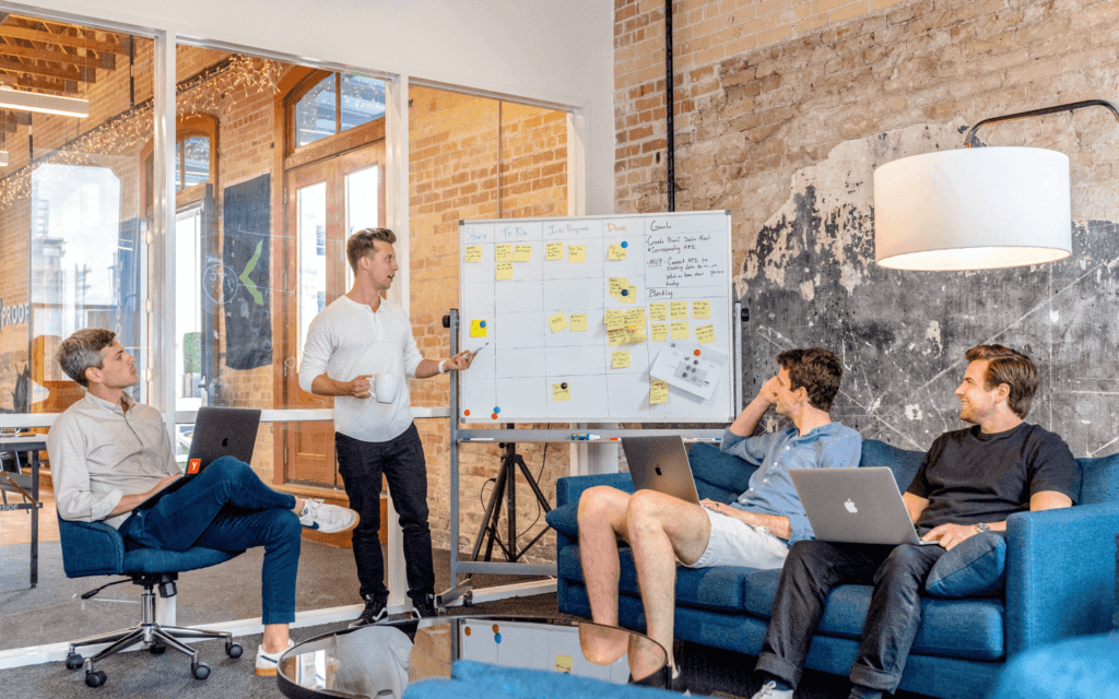 Team of professionals engaged in a Digital Agency Workflow, with one presenting at a whiteboard while others participate from a couch, in a modern brick-walled office.