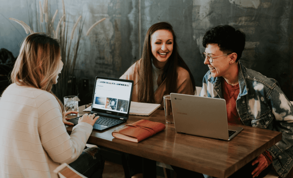 Three people collaborate in a cozy workspace, with one individual browsing a photo sharing site on a laptop, highlighting the social aspect of "How to Caption Images.