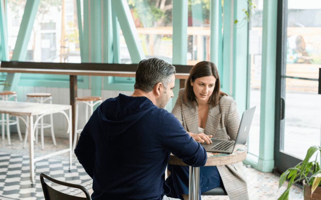 Two professionals engaged in a discussion over a laptop at a café, possibly exploring the 'Best CRM for Digital Marketing Agency' to enhance their marketing strategies and client management.