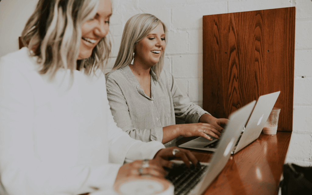 Two happy women working together on a laptop in a well-lit office space, demonstrating successful client retention through teamwork.