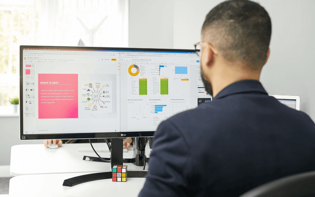 SEO expert reviewing a Keyword Research Report on dual monitors, with one showing an SEO presentation and the other displaying website analytics.