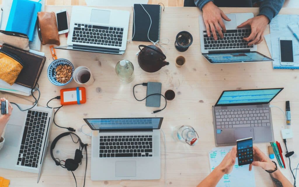 Top view of a busy workspace with multiple laptops displaying graphs, surrounded by mobile devices, notepads, and a teapot, indicating a team analyzing an Email Campaign Report.