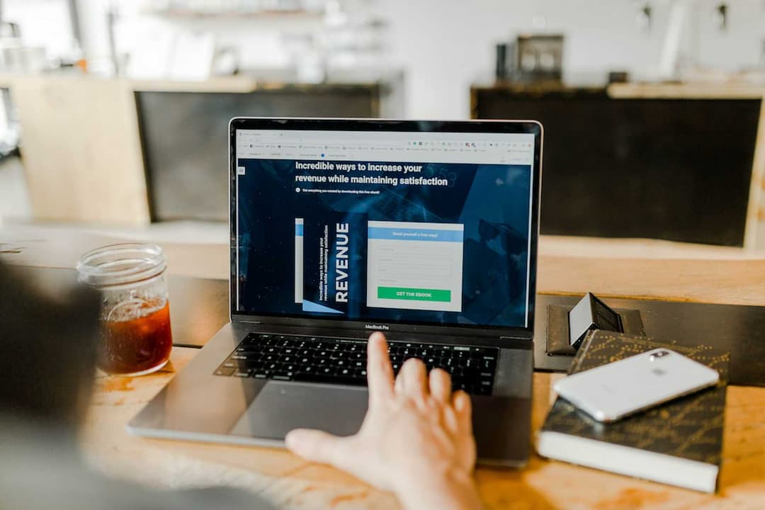 Person using a laptop on a wooden table, viewing a webpage on creating lead magnets to boost revenue and customer satisfaction, with a cold drink and smartphone beside.