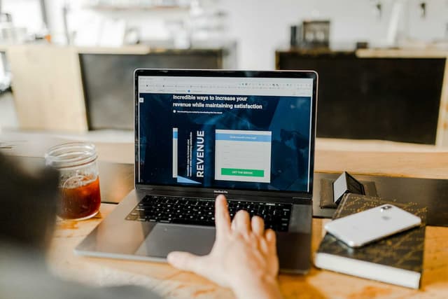 Person using a laptop on a wooden table, viewing a webpage on creating lead magnets to boost revenue and customer satisfaction, with a cold drink and smartphone beside.