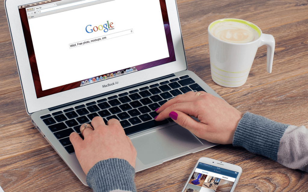 Person using MacBook Air for Google searches, indicative of search engine marketing research, with a smartphone and coffee mug on a wooden surface.