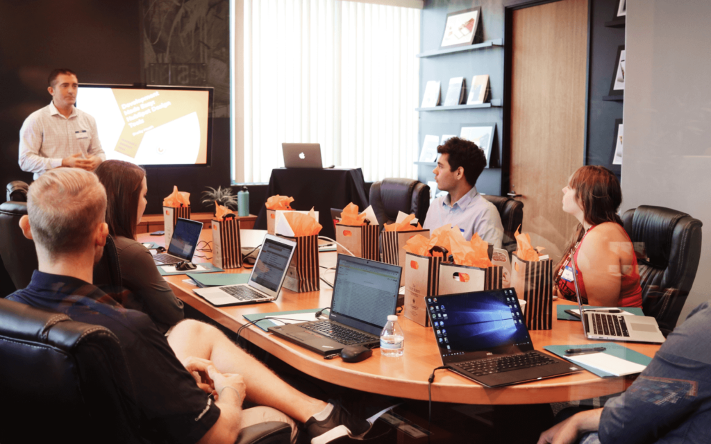 A presenter leading a business meeting on 'Reporting Best Practices' with attentive participants in a modern conference room filled with technological devices.