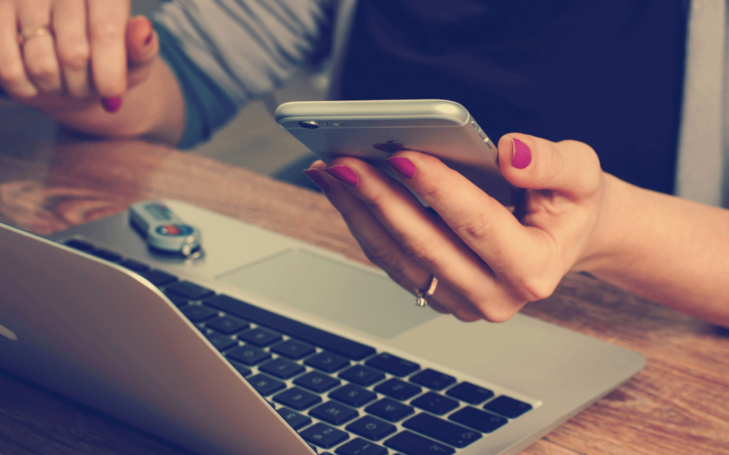 Close-up of a digital marketer's hands using a smartphone, with a laptop and car keys on a wooden table, representing the flexibility and mobility required in the digital marketing industry.