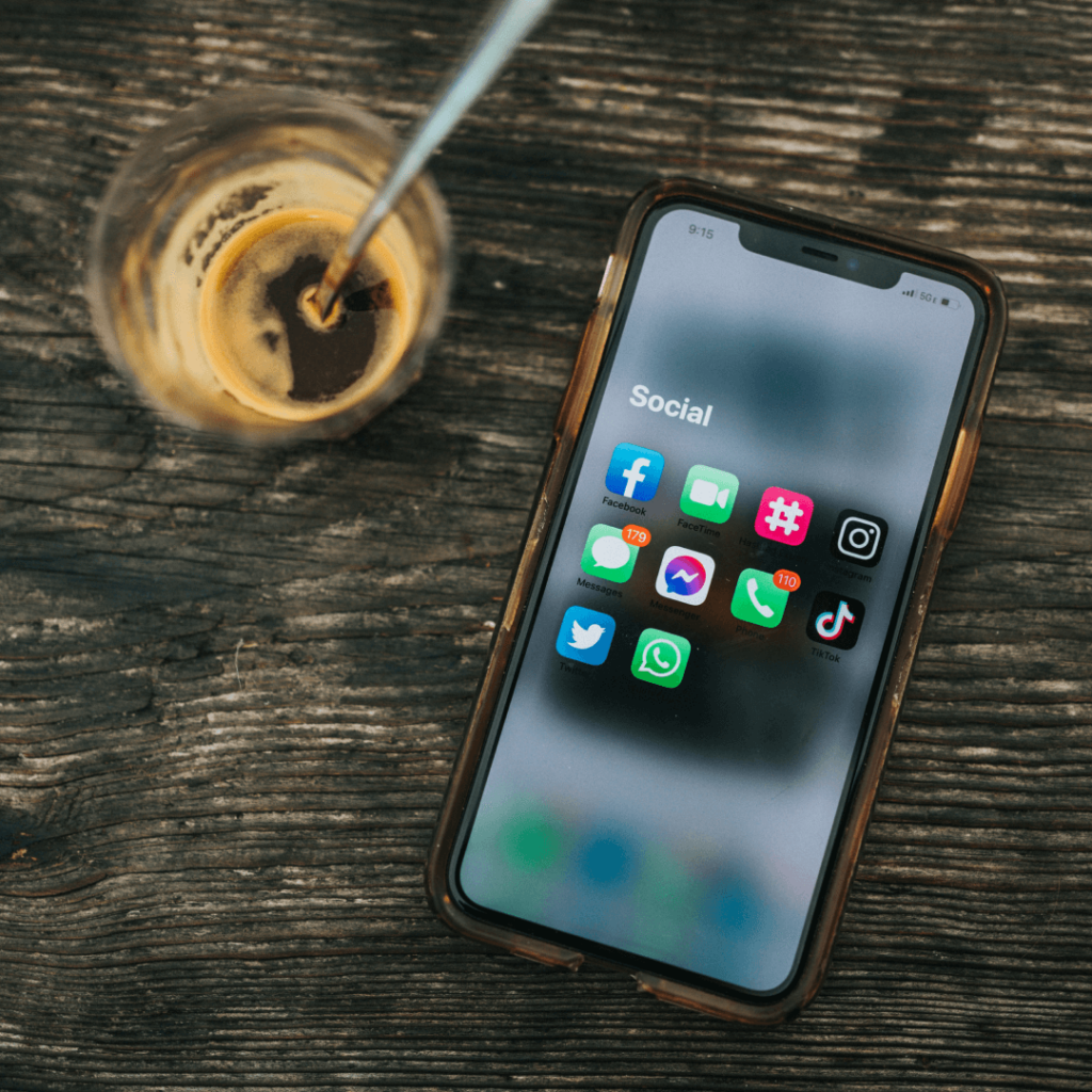 Smartphone with social media apps on display, representing the digital marketing tools used for engagement and strategy on a textured wooden table, with a hint of a coffee break in the background.