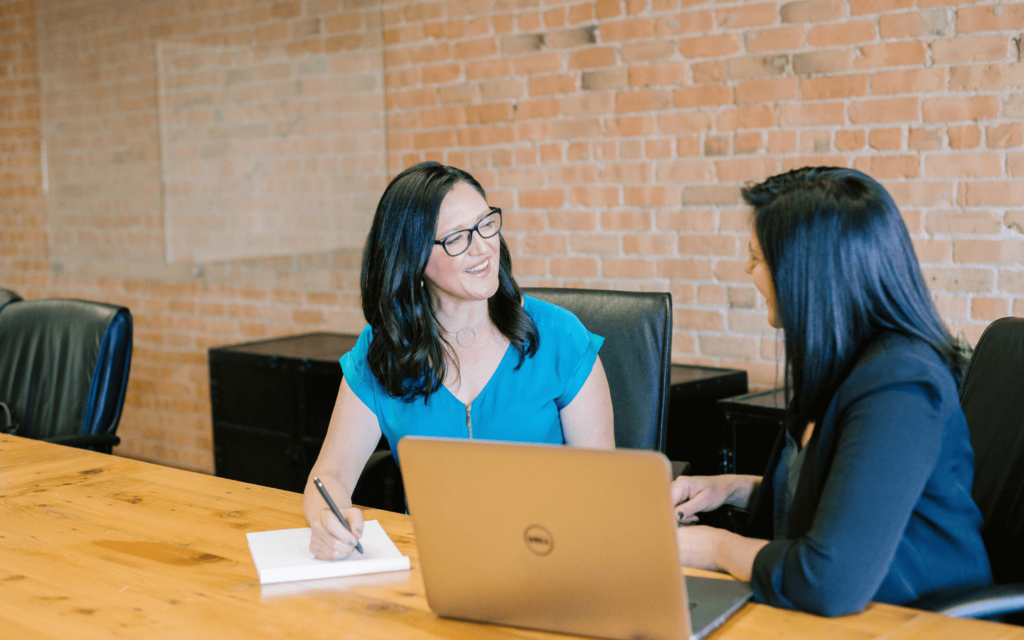 Two women in a professional meeting, with one writing notes and the other using a laptop, possibly demonstrating the use of a White Label SEO Report Generator in their workflow.