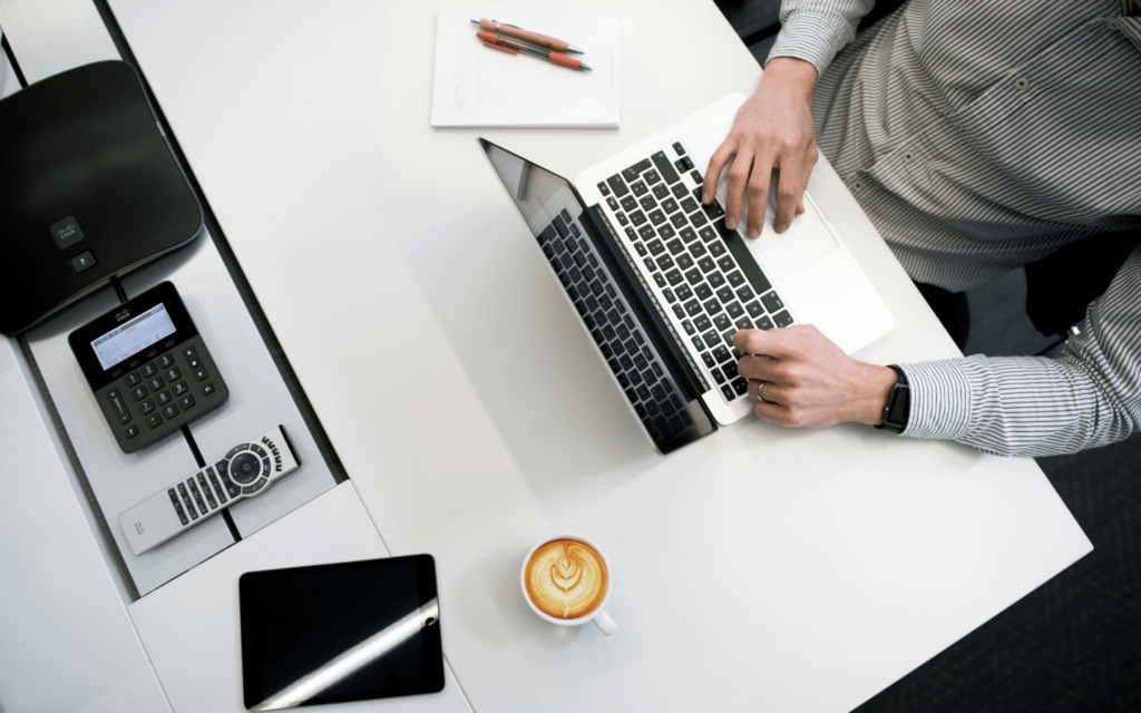 A professional at a minimalist desk working on 'White Label Google Analytics Reports', with a laptop, digital tablet, conference phone, and a cup of coffee, symbolizing efficient and focused analytics work in a contemporary office setting.