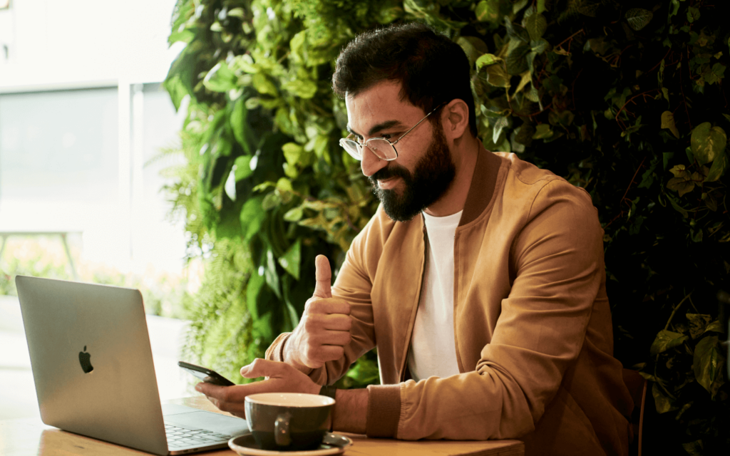 Contemporary marketer working on marketing strategies with a laptop and smartphone in a café surrounded by greenery, giving a thumbs up.