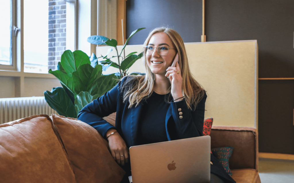 Businesswoman discussing marketing strategies on the phone, seated comfortably with her laptop in a well-lit room with green plants.