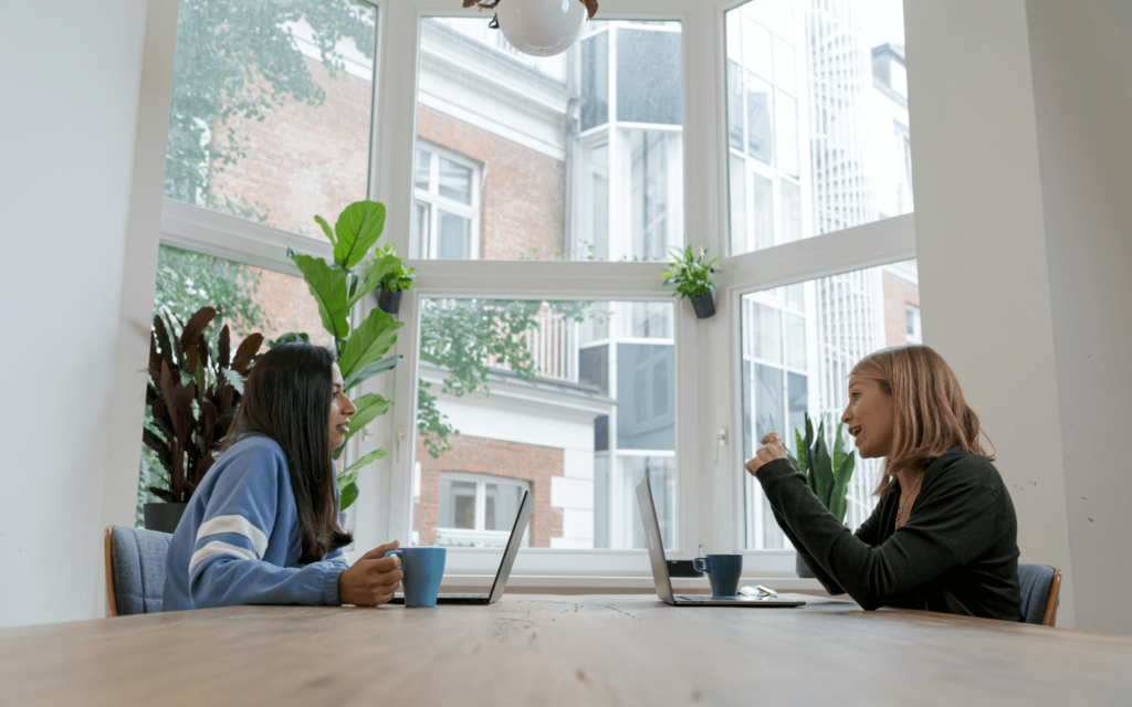 Two professionals in a client meeting, consulting over laptops in a contemporary office filled with natural light and greenery, discussing analytics and strategies using client reporting software.