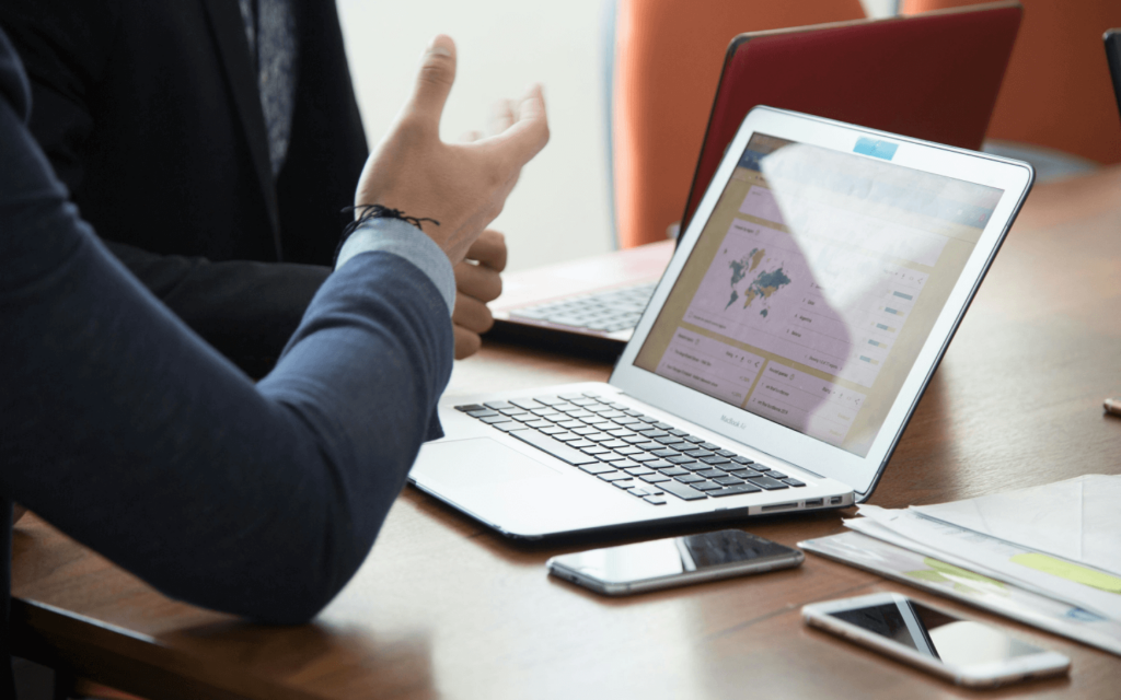 Business professional gesturing during a discussion with a laptop opened to a Client Reporting Software dashboard, indicating an interactive analysis session in a corporate setting.