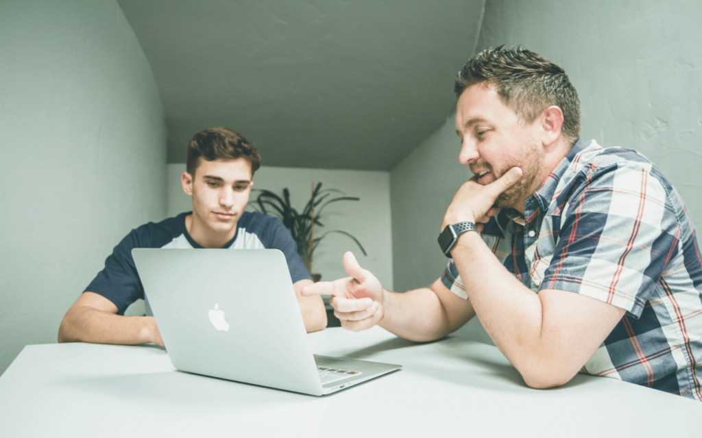 Two professionals in a casual discussion, using a laptop presumably displaying Client Reporting Software to analyze client data, embodying a laid-back yet focused work culture.