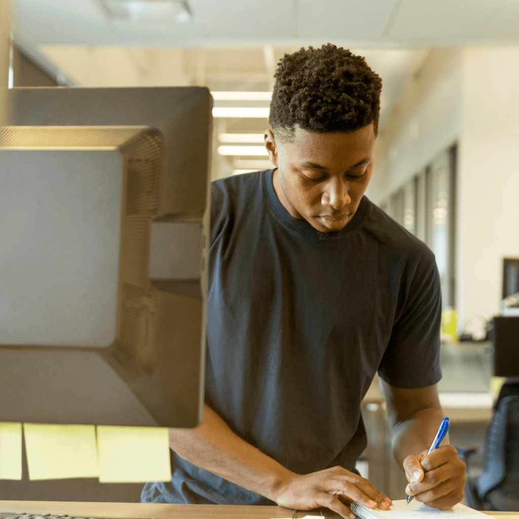 A concentrated individual writing notes beside a desktop computer, potentially reviewing data from client reporting software, with sticky notes attached to the screen for organization.