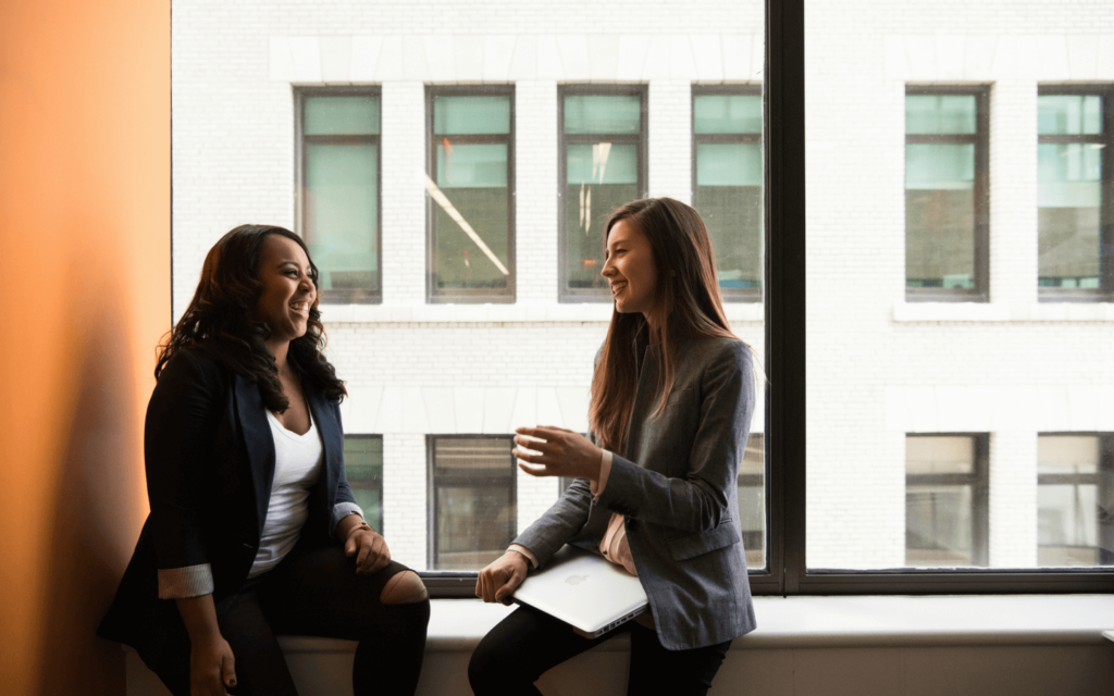 Two smiling women discussing a website SEO report in an office setting, one holding a laptop, symbolizing a collaborative approach to SEO strategy.