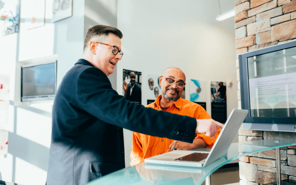 Two men in a professional setting, with one pointing at a laptop screen, possibly discussing a website SEO report, highlighting a collaborative effort in SEO strategy and analysis.