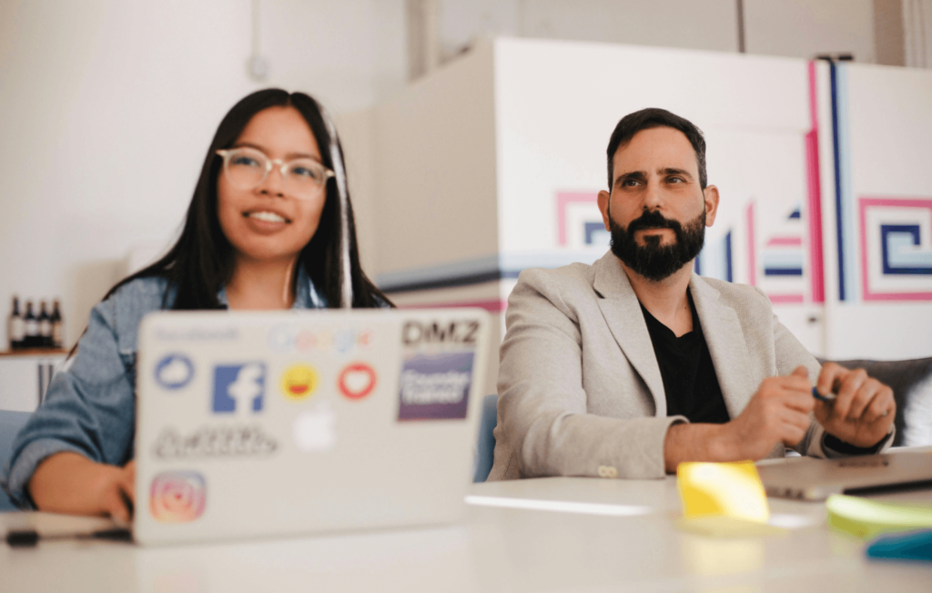 Digital marketing professionals in a discussion, with a focus on a laptop featuring social media stickers, indicative of a 'White Label SEO Reporting Dashboard' planning session.