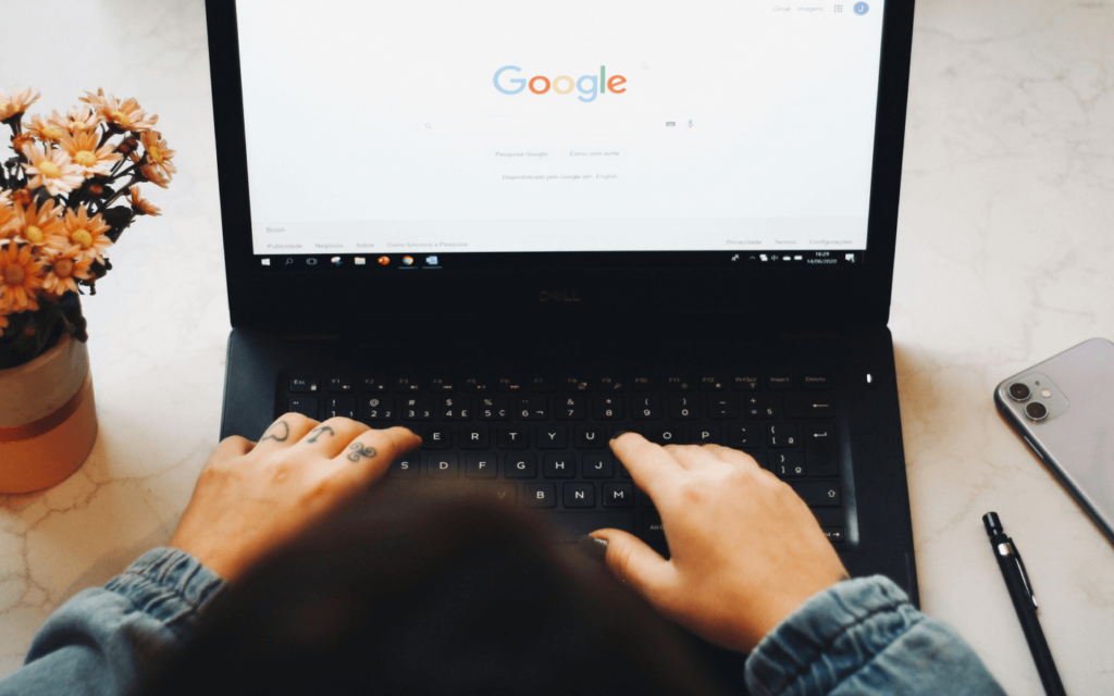 A user with tattooed hands typing on a laptop with a Google search homepage on the screen, possibly researching GA 4 reports.